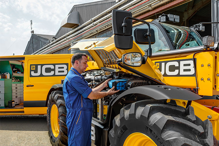 Mies huoltaa JCB-teleskooppikuormaajaa, takana JCB-logolla varustettu pakettiauto sekä maatilan rakennus.